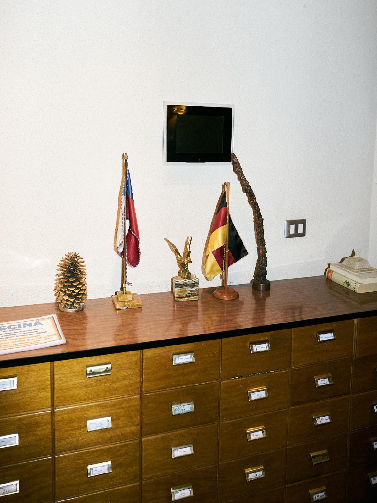 𝐈𝐈. The brief glimpse of a settler's dream - Chilean and German flags behind the desk in the...