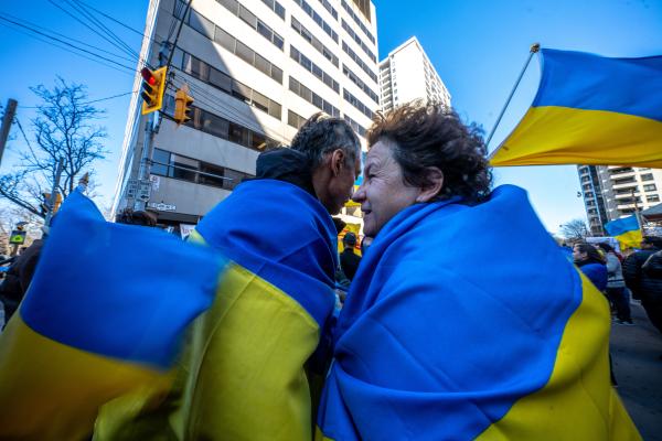 Image from [Ongoing] Ukrainian Toronto Protests - Two people wearing Ukrainian flags embrace. People gather...