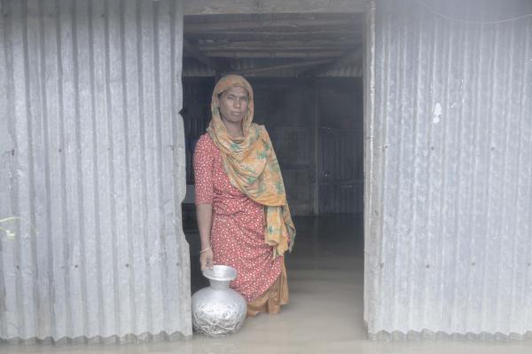 Image from Flood Survival People -  Hena Begum stands in front of her flooded house with a...