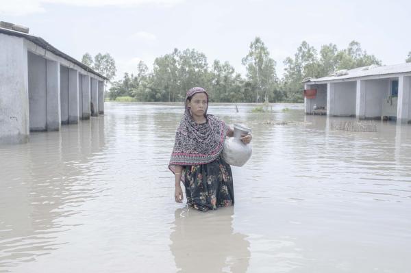 Image from Flood Survival People -  Manjura Begum walks through floodwaters carrying a jar...