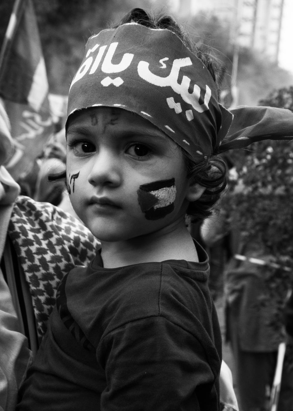 From the river to the sea - A child with a Palestinian flag painted on her face and a bandana which translates &quot;Here...