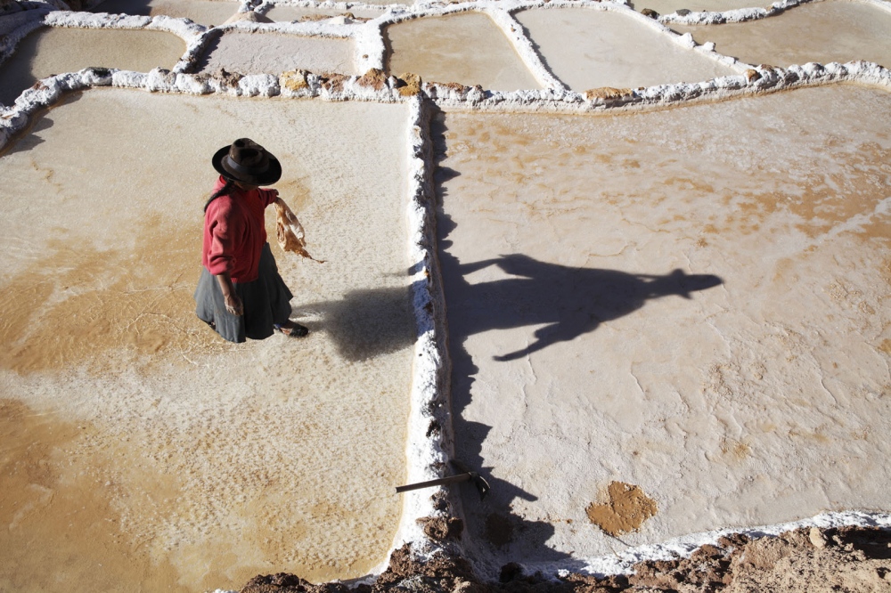 A Quechuan lady walks across the ancient salt pans on Salinas de Maras casting a long shadow. Built by the Inca&#39;s this UNESCO status site is still used to produce salt by members of the Maras community. Maras, Peru.
