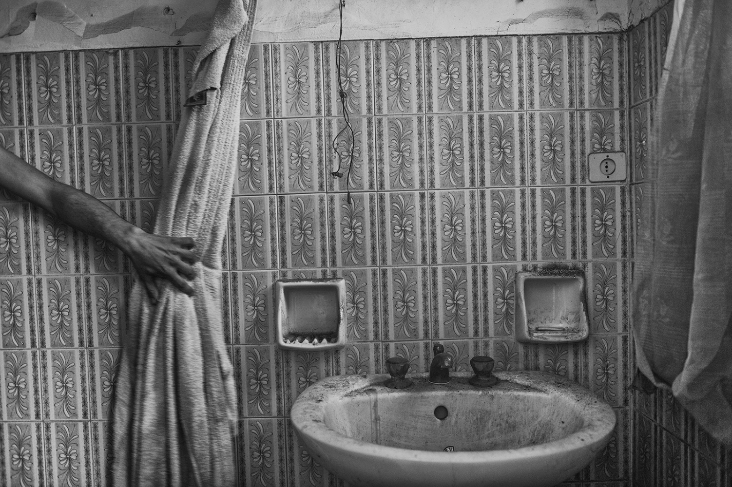  The hand of a man moves a dirty and ragged bathrobe to show the washbasin of a bathroom covered with a layer of dirt. 