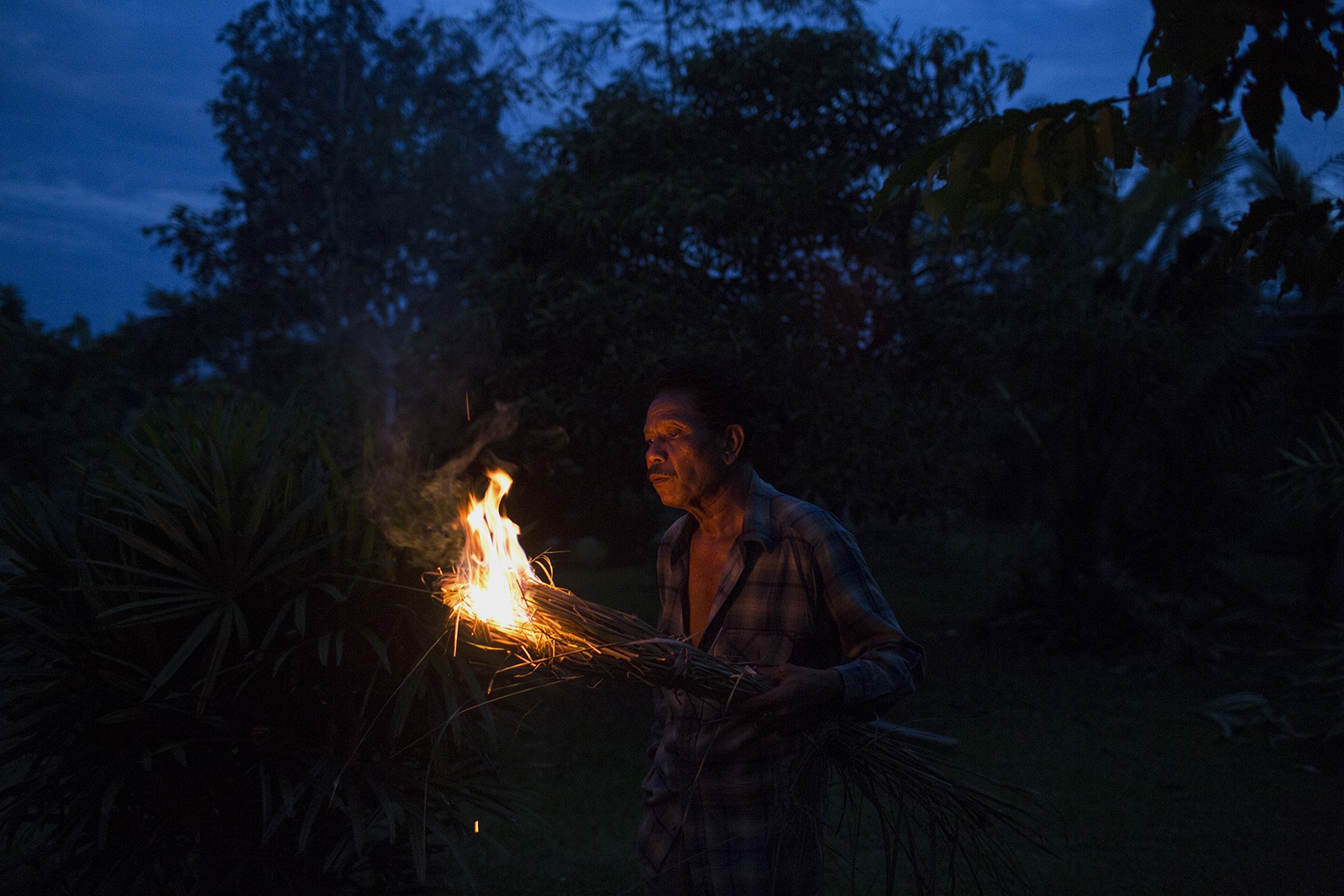 A VILLAGE UNDER SIEGE - Pratip Rakhungthong, 61, lights dried lemongrass grown in...
