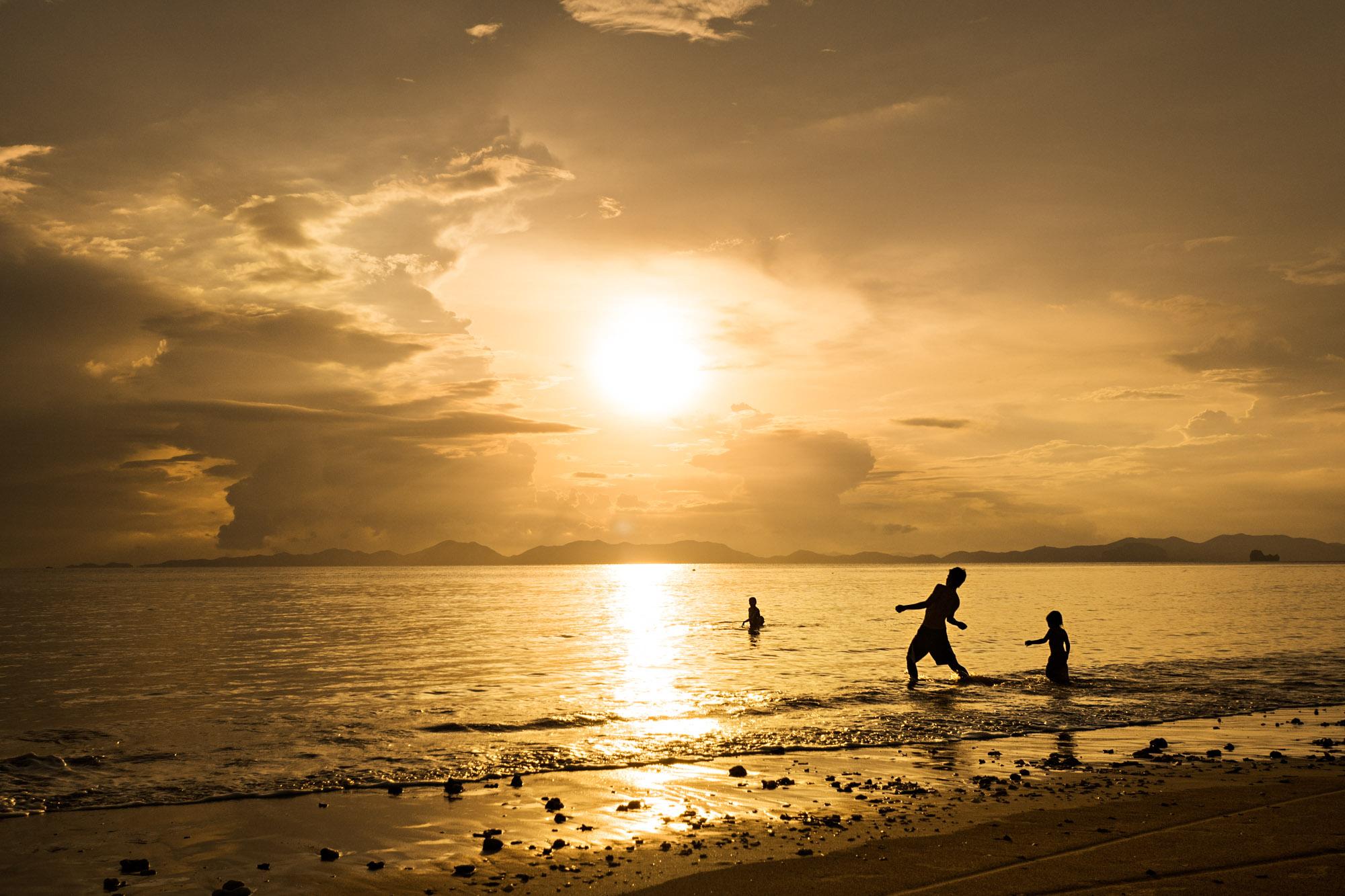 Travel - A father plays with his children in the ocean at sunset...