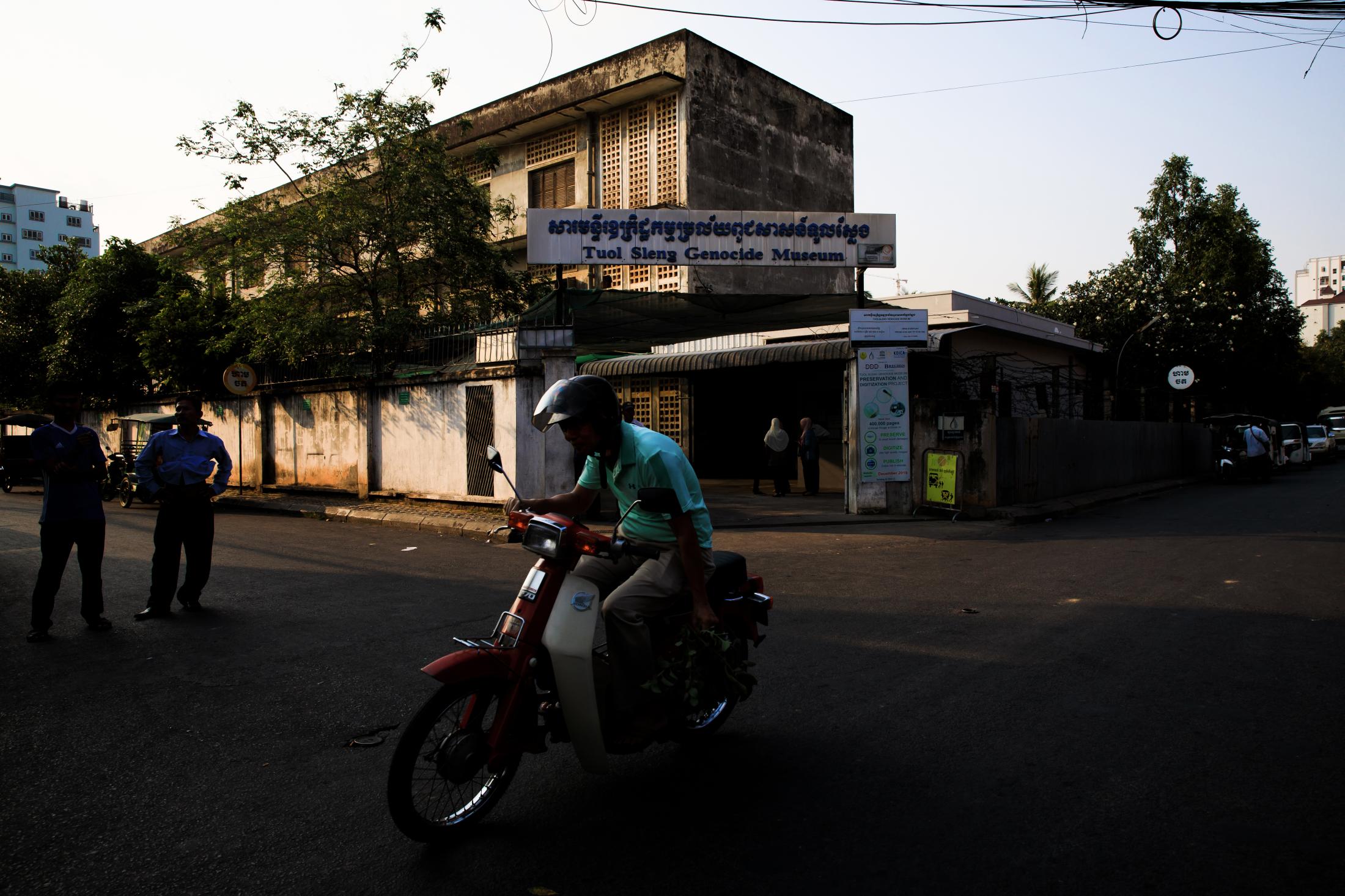 3Y8M20D -  A man drives his motorcycle in front of Tuol Sleng...