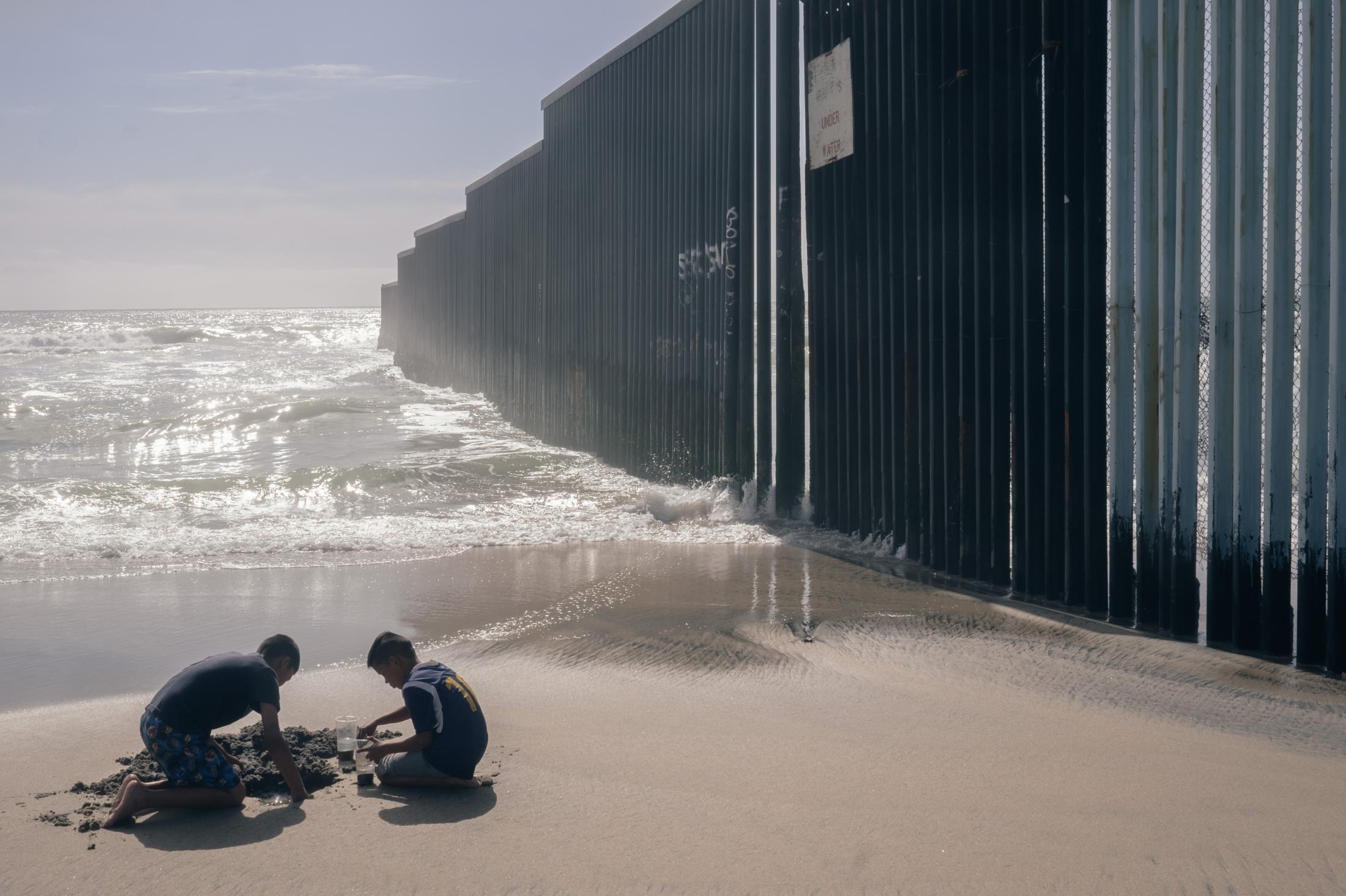 Southern Border - Two children play in the sand next to the wall that...