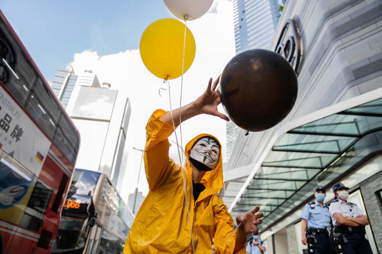 A protestor dressed in a yellow raincoat with black, yellow, and white balloons. Two years ago, a man named Marco Leung Ling-kit fell to his death from scaffolding of a nearby building during social unrest in Hong Kong. Dozens of people laid white flowers, candles, and ribbons in mourning.