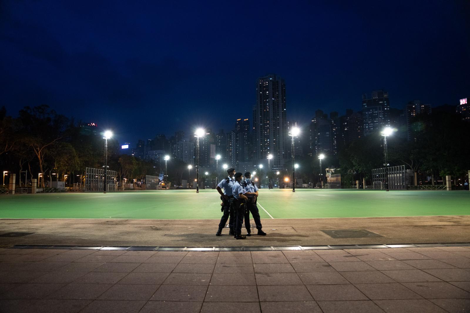 Three police officers huddle together in a blocked off and empty Victoria Park, Hong Kong on June 4, 2021. For the second time since 1989, a Tiananmen Square vigil was banned in Hong Kong, citing concerns of public health safety. Despite Victoria Park (the typical meeting location of the vigil) being closed with 3,000 police officers deployed around the area, Hong Kong people still showed up near the park, with the flashlights on their phones turned on. By the end of the night, at least six people would be arrested and twelve people fined.