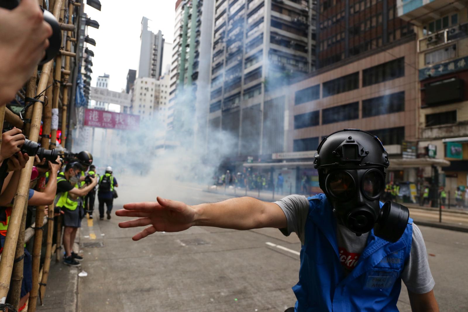 A member of the police media liaison pushes back journalists as tear gas is fired in the background. On the first day of the passing of the National Security Law, protests erupted across Hong Kong. In 2020, the ranking of press freedom in Hong Kong dropped to the 80th place, with increasing pressure and constrictions on media.