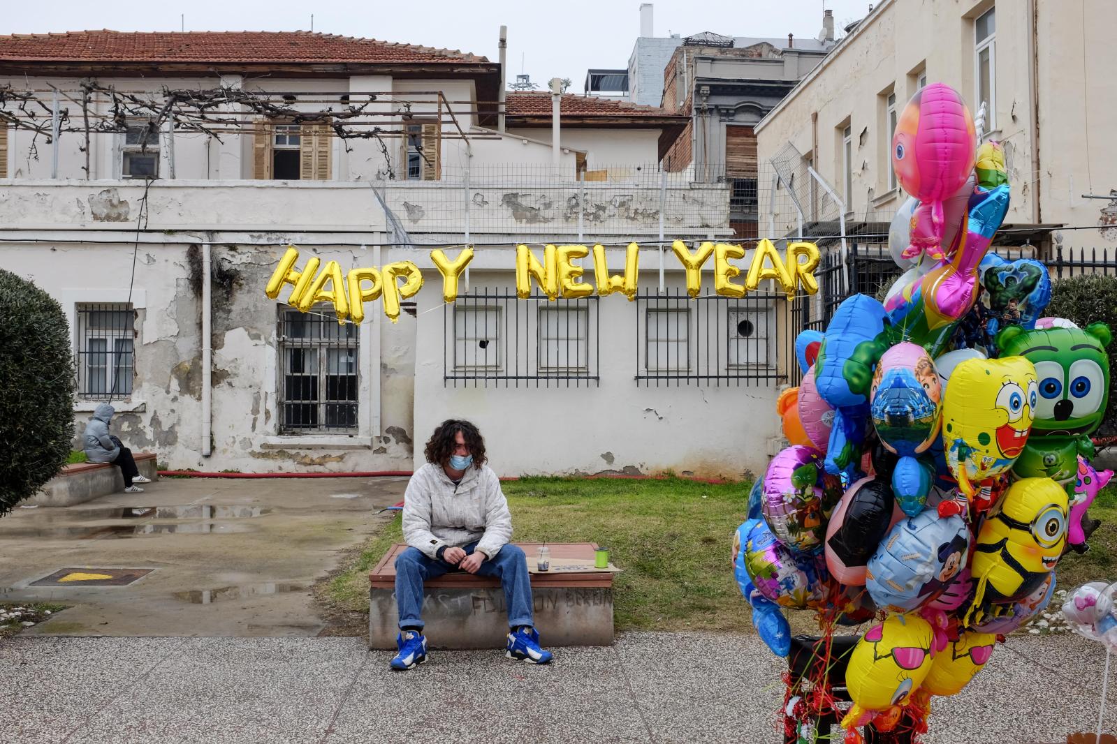 A man wearing a face mask sits on a bench next to balloons, some of which read &quot;Happy New Year&quot;. Around Christmas time, amid rapidly rising Coronavirus infections, the Greek government imposed a strict mask mandate in all public places, including the outdoors. Thessaloniki, late December 2021.